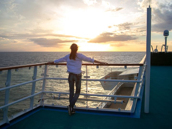 A woman stands facing away on a cruise ship deck looking at the sunset with her arms spread across the railing.