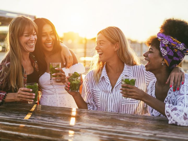 A group of women sitting outside together laughing while all holding drinks.