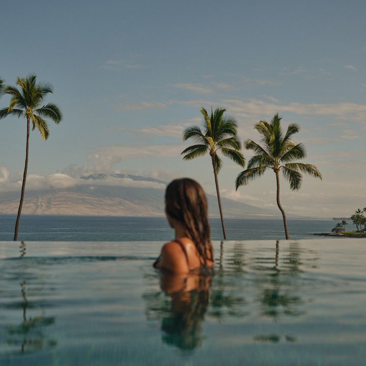A woman seen from the back of her head at the edge of the infinity pool at the Four Seasons Resort Maui looking at the ocean and mountain views.