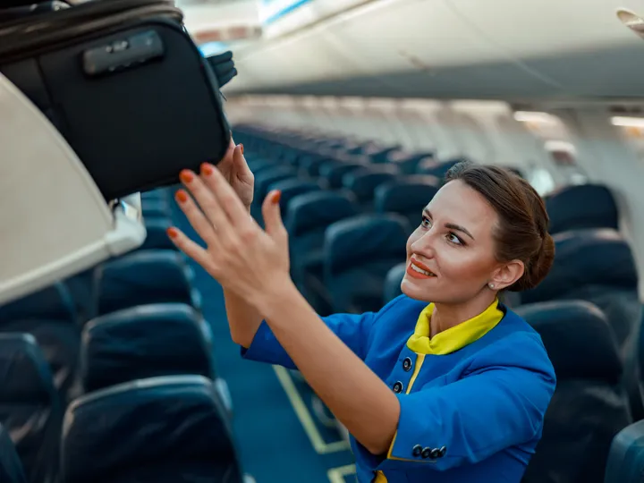 A flight attendant loads a suitcase into an overhead compartment.