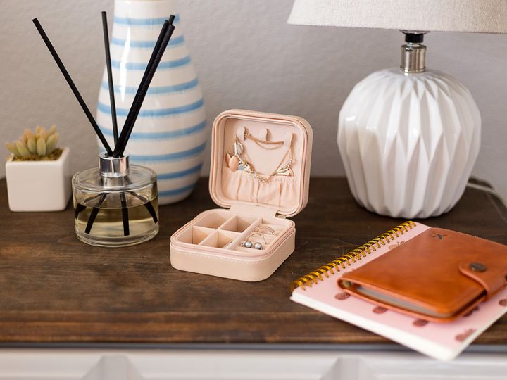 A small compact jewelry case sitting on a dresser next to a lamp, notebook, aromatherapy sticks, and a succulent.