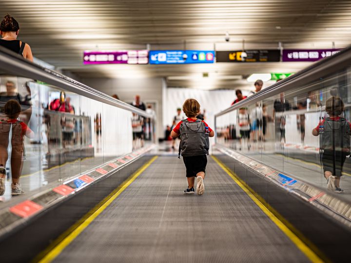A small child wearing a backpack runs down a moving walkway in an airport.