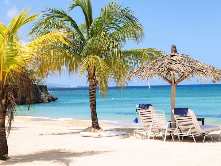 An empty tropical beach with white sand, swaying palm trees, blue water, and two empty lounge chairs.