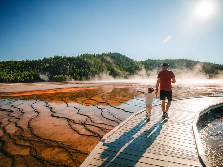 A man and his child walk on a boardwalk next to geysers at Yellowstone National Park.
