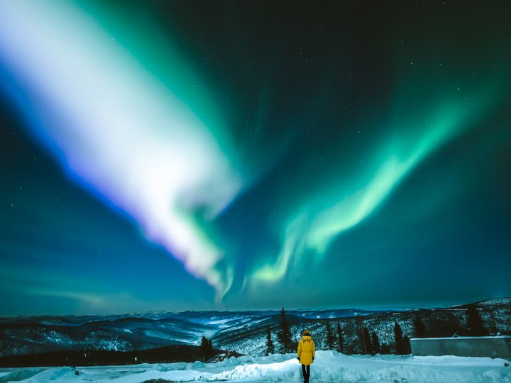A person in a yellow coat stands on a snowy landscape in Fairbanks, Alaska as neon blue and green northern lights appear in the night sky.