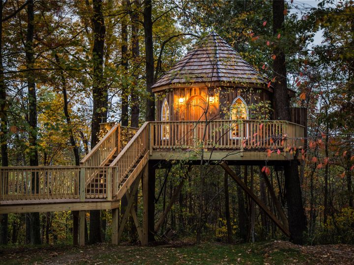 A cylindrical-style treehouse with a shingled roof and stairs leading up to it is perched in the woods.