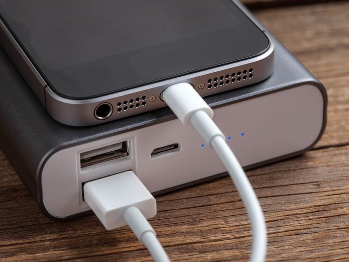 A power bank is charging an iPhone with it stacked on top of it, connected by a cord, sitting on a wood surface.