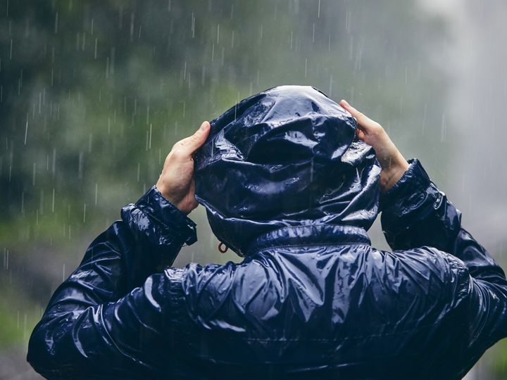 A person stands in the pouring rain, seen from behind, holding their hood up.