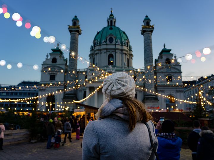 A woman is seen from behind, standing at the Ard Ardvent Christmas market in Vienna with string lights and booths in front of a church.