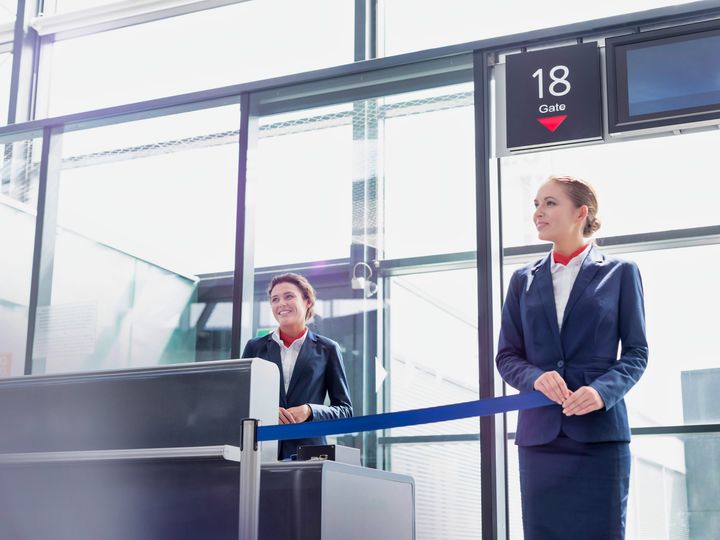 An airline gate agent stands at the gate door with tape partially closing it while another one stands at the desk.