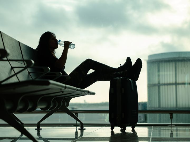 A low angle shot of a woman in dark light at the airport drinking a bottle of water while waiting for her flight with her feet propped up on her suitcase.