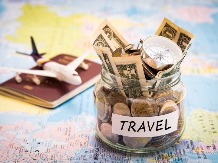 A toy plane sitting on top of a passport next to a jar filled with change and dollars labeled travel, all on top of a map.