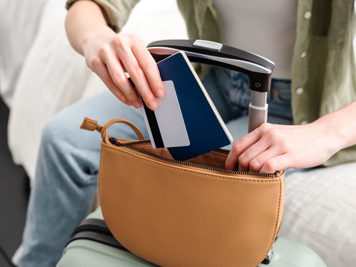 A person is seen without the face visible putting a passport and credit card into a small purse on top of luggage.
