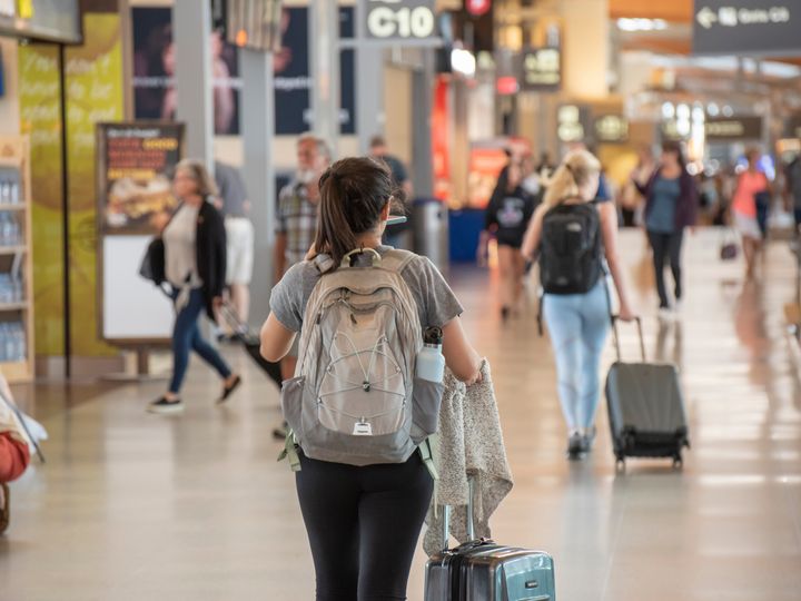 A woman is seen from behind wearing a backpack and pulling her suitcase while walking next to airport shops.