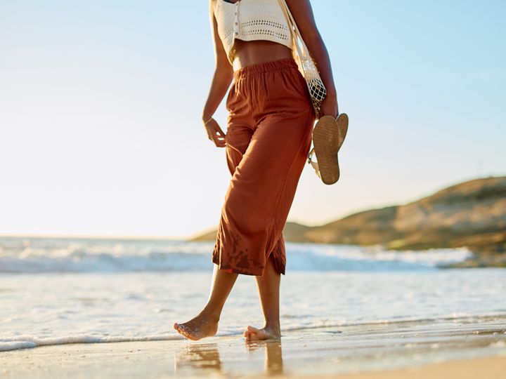 A woman is seen from the waist down walking on a tropical beach in the surf carrying her flip flop sandals.