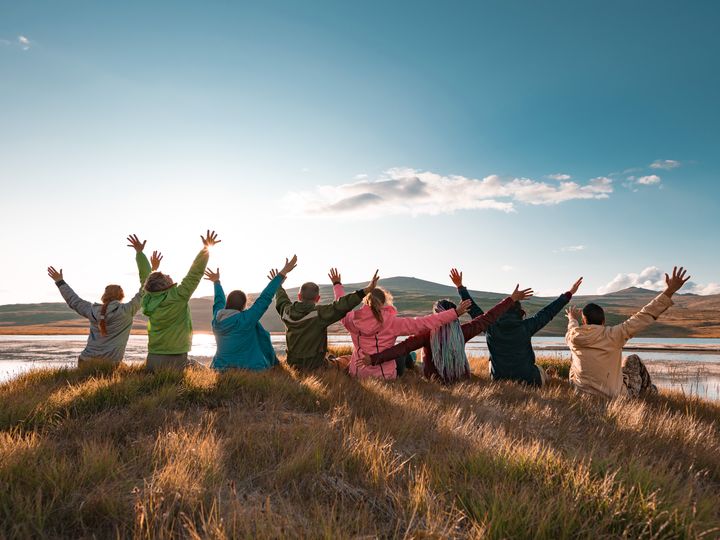 A group of people sits with their backs to the camera and arms in the air in a scenic setting at sunset.
