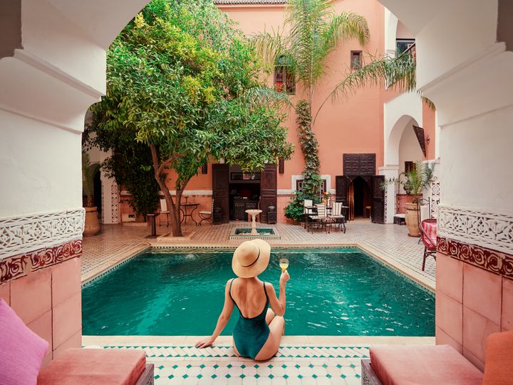 A woman wearing a hat and holding a glass of wine next to an interior Moroccan-style courtyard pool.