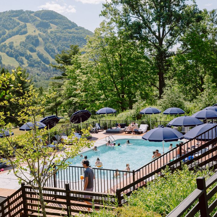 Stairs lead down to a pool surrounded by chairs and blue umbrellas next to green mountains.