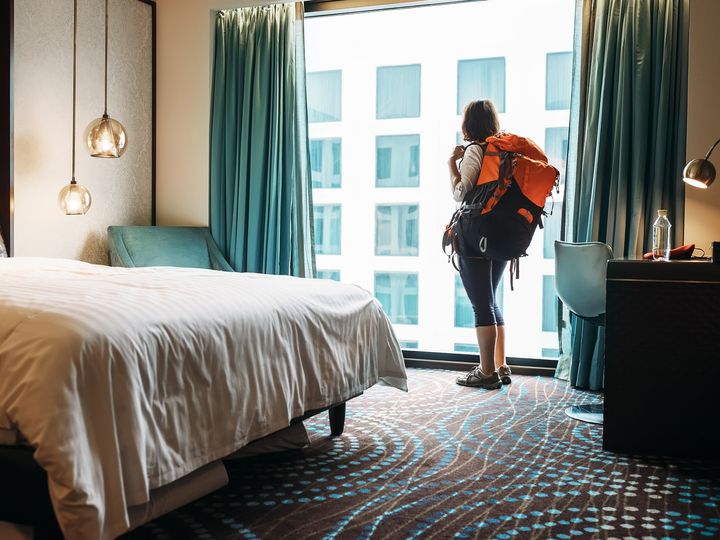A woman stands at a window wearing a large backpack in a hotel room next to the bed.