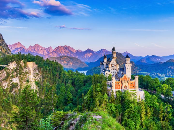 The famous Neuschwanstein Castle in Bavaria, Germany, surrounded by rolling hills and mountains, as well as green trees and a lake in the distance.