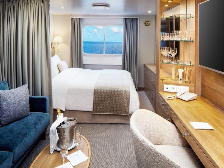 A cruise cabin with a bed and a curtain separating a living area with a couch, table, and desk, in a windowed stateroom.
