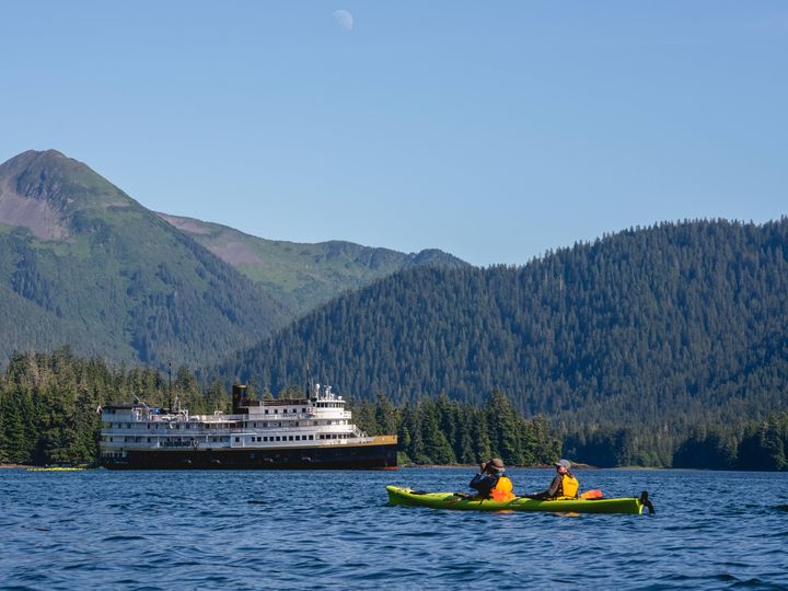 A kayak is seen on the water in front of an expedition cruise boat in a mountainous area.