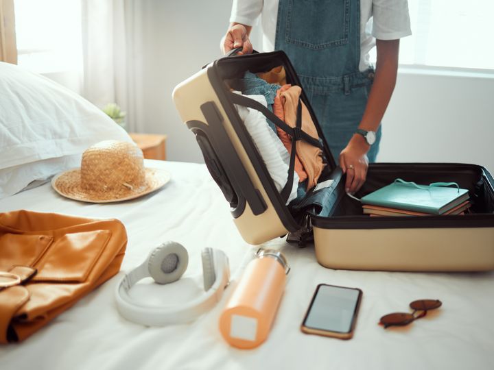 Different travel supplies laid out on a bed with a person packing an open suitcase.
