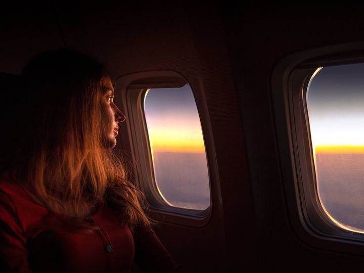 A woman rests her head against the airplane window while looking out at the sunrise.