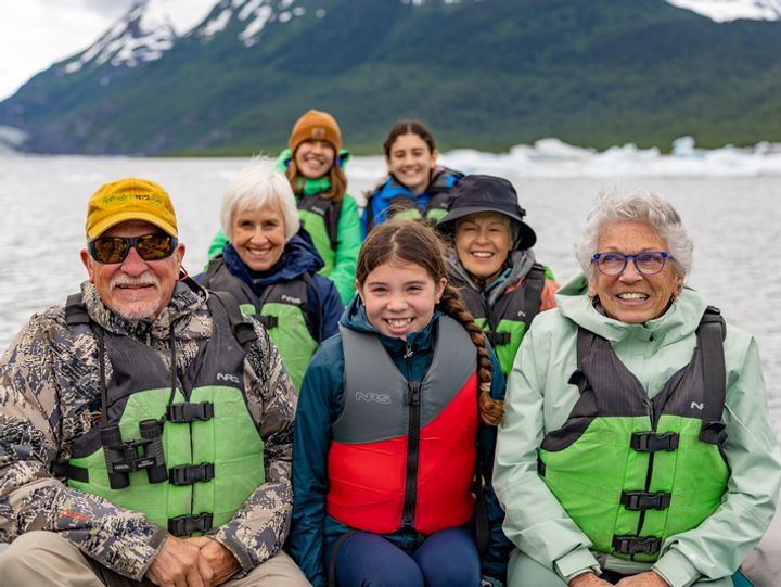 A group smiles for a photo in a boat next to snow-capped mountains with young children and senior-age grandparents.