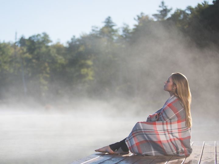 A woman sits on a dock at the water’s edge as steam rises off it, wrapped in a blanket.