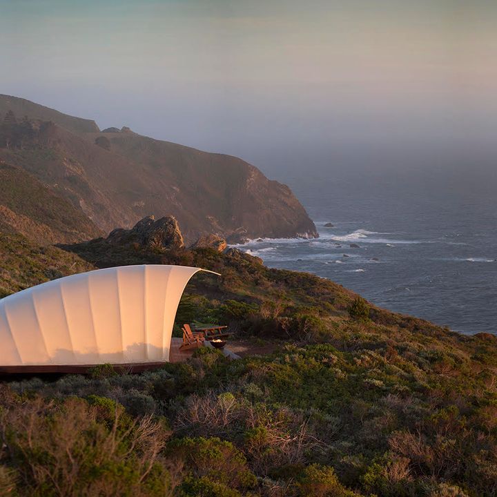 A large white tent with a deck with chairs facing the ocean surrounded by the natural beauty of Big Sur at sunset.