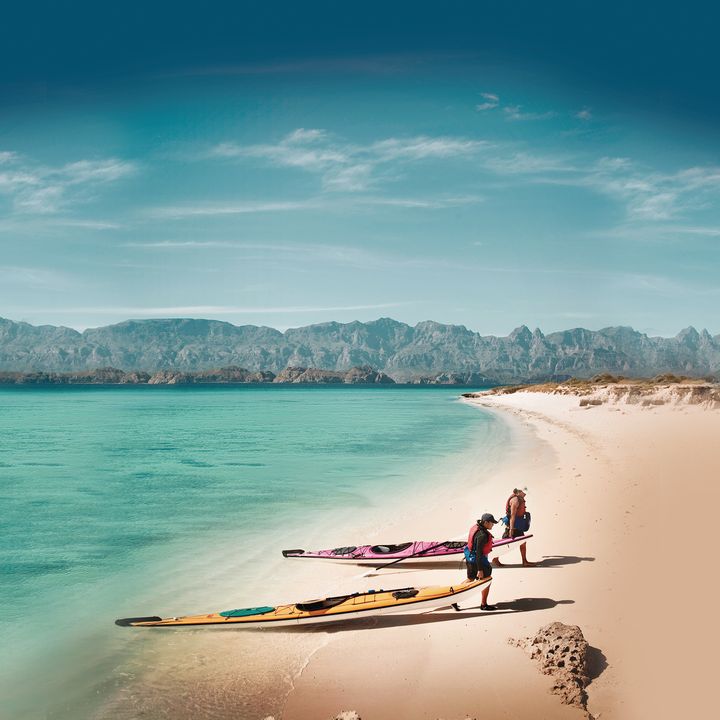 Two people drag their kayaks out of the water on a beautiful beach that has no crowds, white sand, turquoise water, and mountains in the distance.