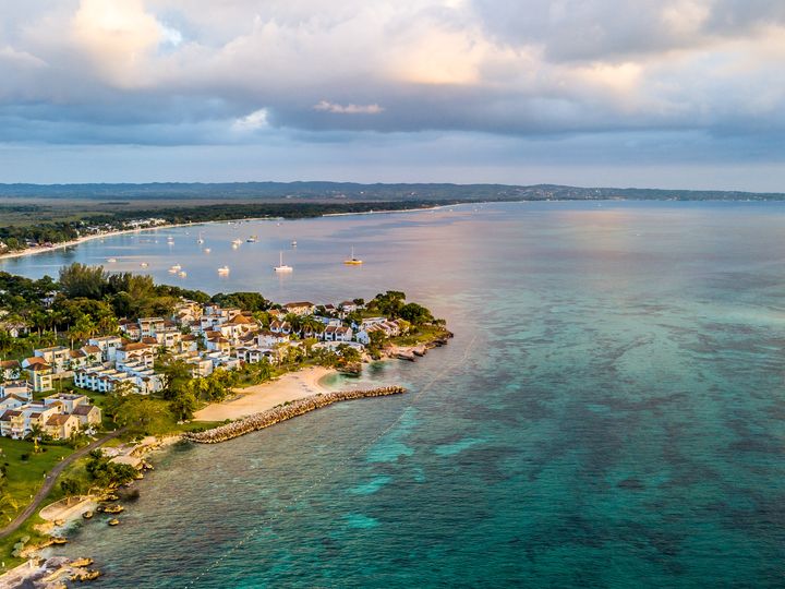 An aerial view over the coastline of Negril, Jamaica with buildings along the shore, boats dotting the water, and beautiful turquoise water.
