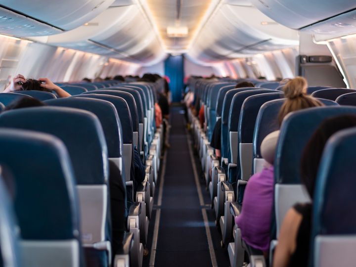 A view from the back of a plane looking forward through the aisle with rows of plane seats filled with passengers.