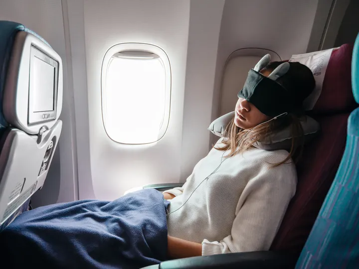 A woman sleeping on a plane while wearing an eye mask next to a window.