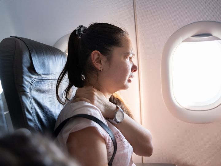 A woman sits at the window seat on the plane holding her neck and making a face in pain.