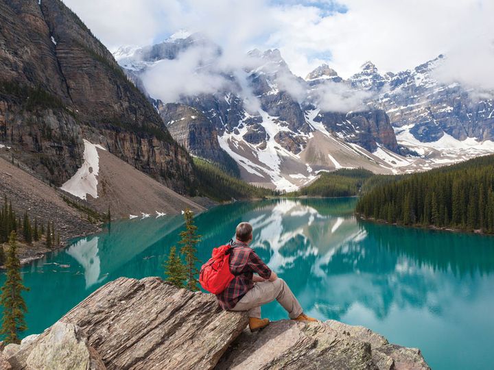 A man with a backpack sits on a rock overlooking snowcapped mountains and a blue lake.
