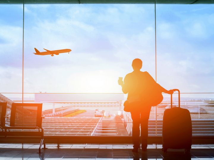 A person is seen looking through an airport window holding luggage and a coffee as light shines through.