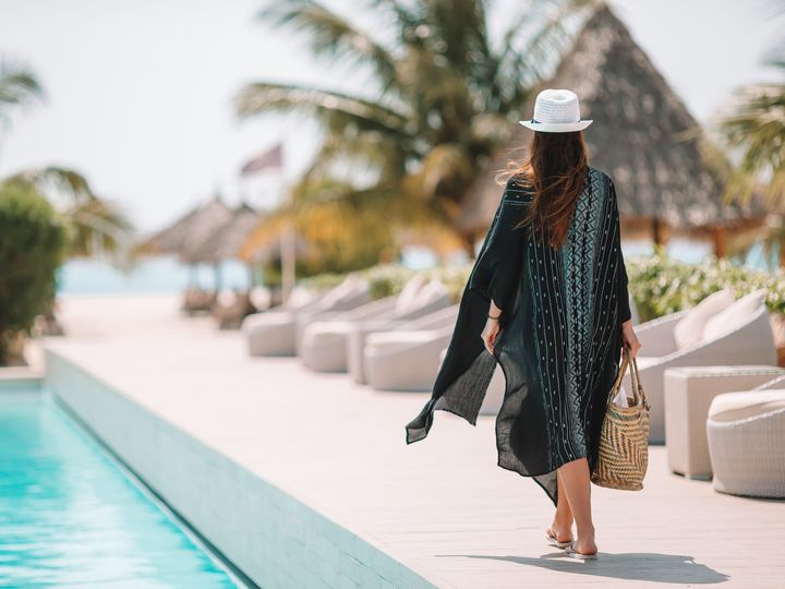 A woman is seen walking by the pool in a caftan and a hat in a luxury hotel resort in a tropical location.