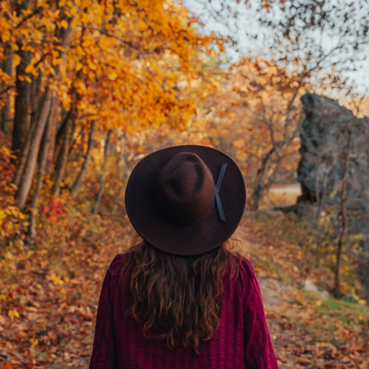 A woman wearing a hat is seen from behind standing in an area amid colorful fall foliage on either side.