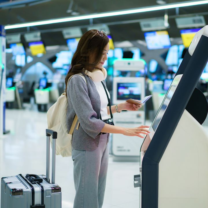 A woman with headphones around her neck and a suitcase at her side stands and operates an airport check-in kiosk