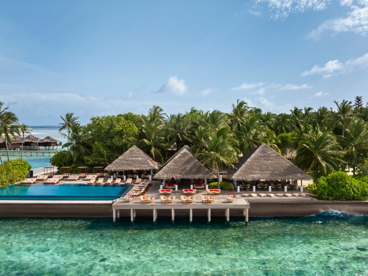 A resort area with thatched roofs and an infinity pool surrounded by palm trees on a platform over the water at Anantara Veli Maldives Resort.