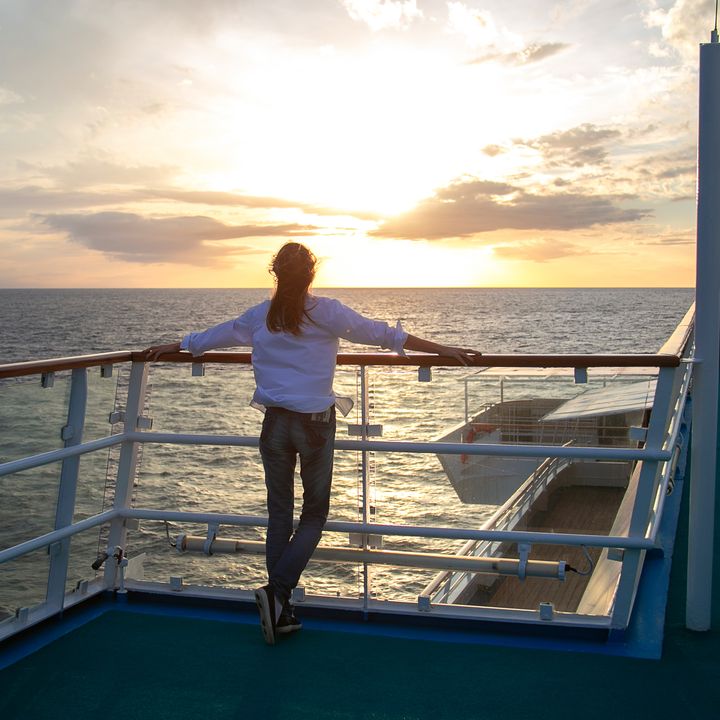 A woman stands facing away on a cruise ship deck looking at the sunset with her arms spread across the railing.