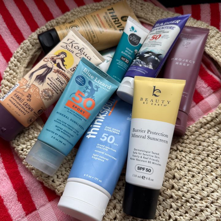 A collection of mineral sunscreen bottles arranged on top of a striped beach towel and a straw beach bag.