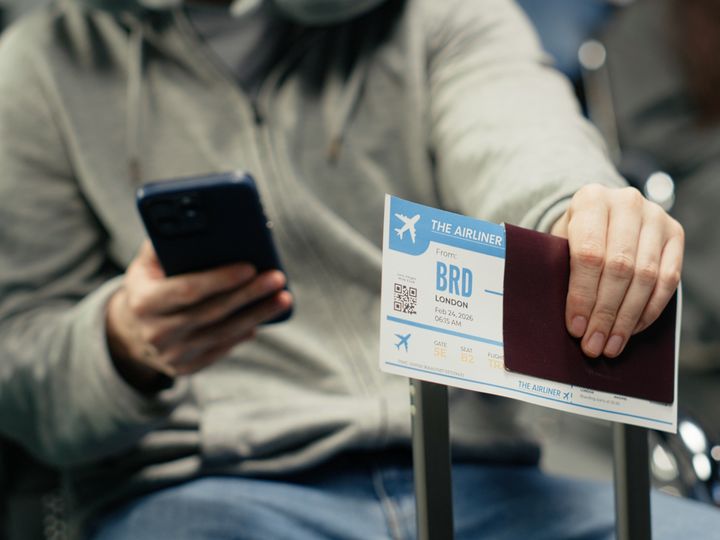 A man sitting at the airport visible from the neck down holding a phone in one hand and a passport and ticket in the other leaning on this suitcase handle.
