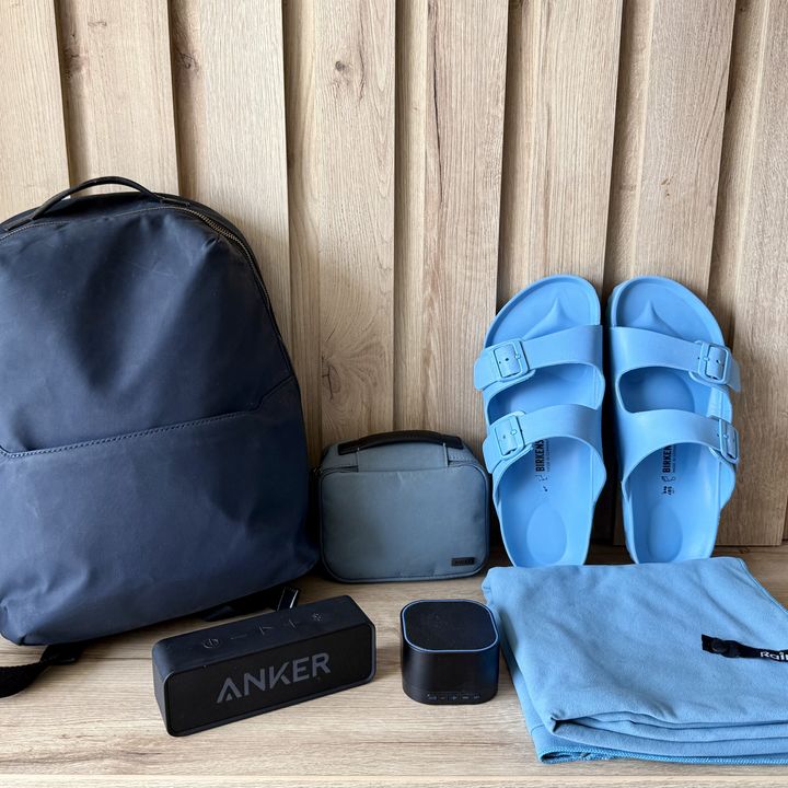 A backpack sits next to a travel pouch, a white noise machine, speaker, blue shoes, and a towel in front of a wood wall on wood floors.