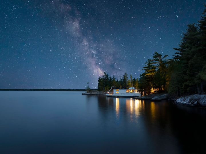 A houseboat moored at the edge of a lake and forest beneath a starry night sky.