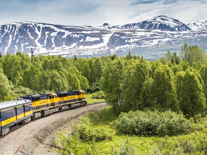 A yellow train labeled "Alaska" passes along tracks next to snow-covered mountains.