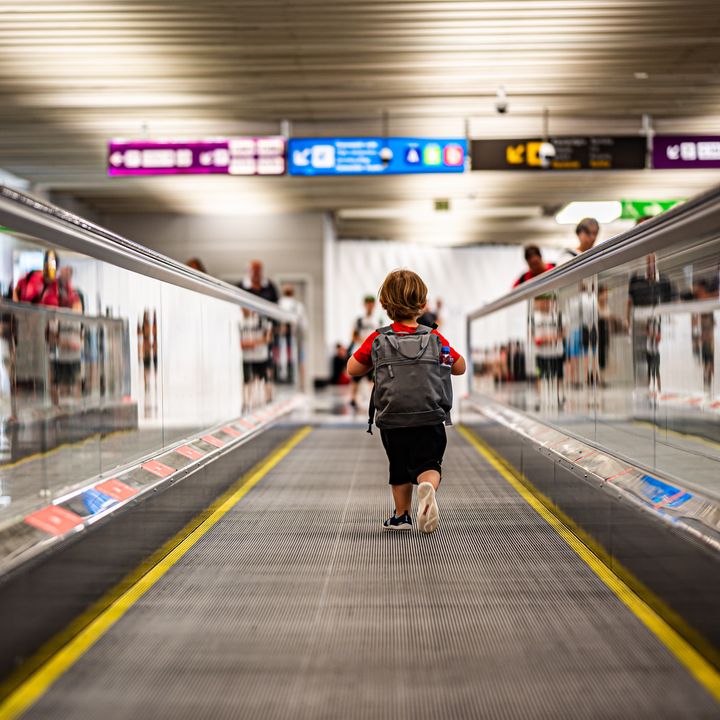 A small child wearing a backpack runs down a moving walkway in an airport.