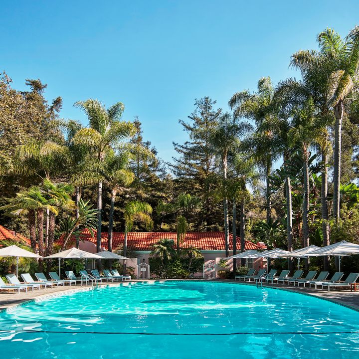 A round circular pool surrounded by pool chairs with a Mediterranean-style building behind it and towering green trees all around.